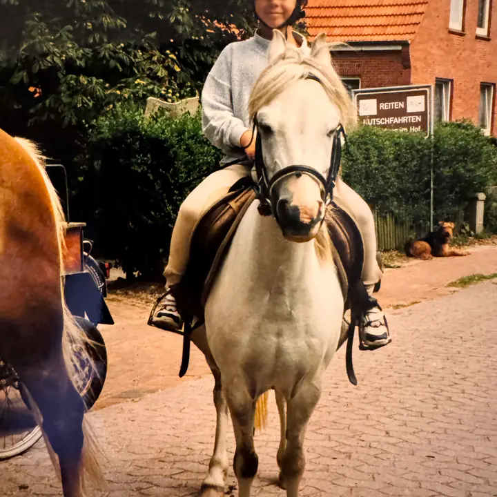 Kind mit Reithelm auf einem hellen Pony vor einem Reiterhof